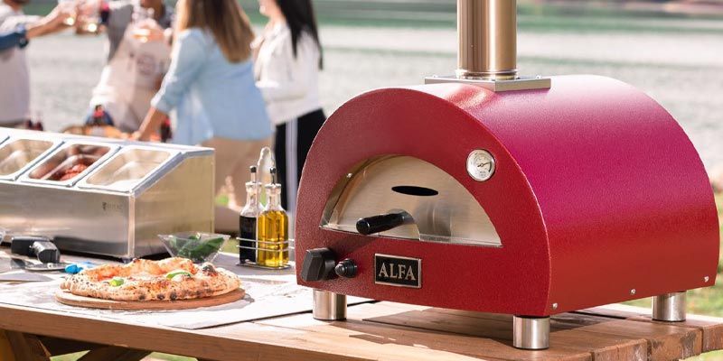 Red outdoor pizza oven with a pizza on a table at a food stall, people blurred in the background