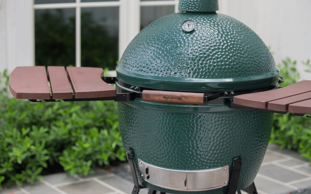 Green ceramic grill with wooden side shelves on a patio, in front of a window and shrubs