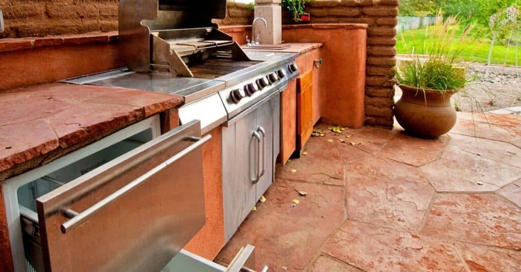 Outdoor stainless steel grill and oven built into a red stone patio kitchen with a planter nearby