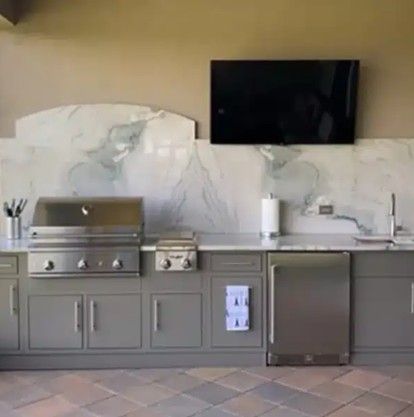 Outdoor kitchen with gray cabinets, stainless grill, sink, refrigerator, and wall-mounted TV.