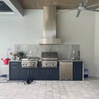 Modern outdoor kitchen with stainless steel grill, sink, and dark cabinets against a marble backsplash