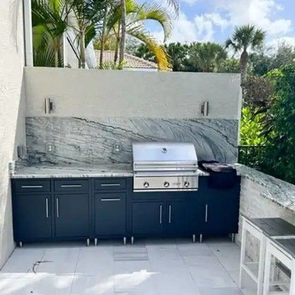 Outdoor patio with built-in black cabinets, stainless grill, and stone backsplash beside lush greenery
