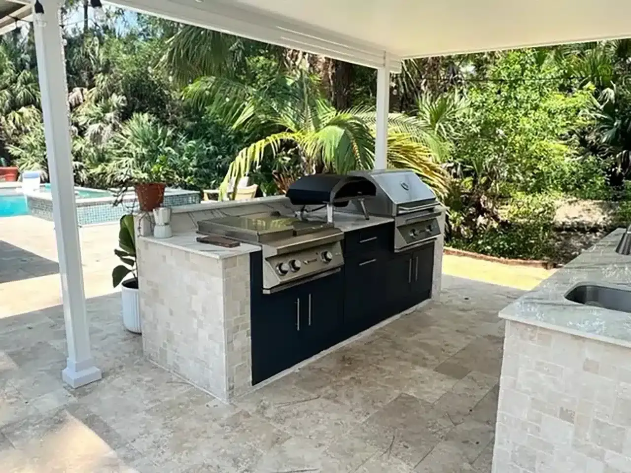 Covered outdoor patio with built-in stone grill, sink, and countertop, surrounded by tropical greenery
