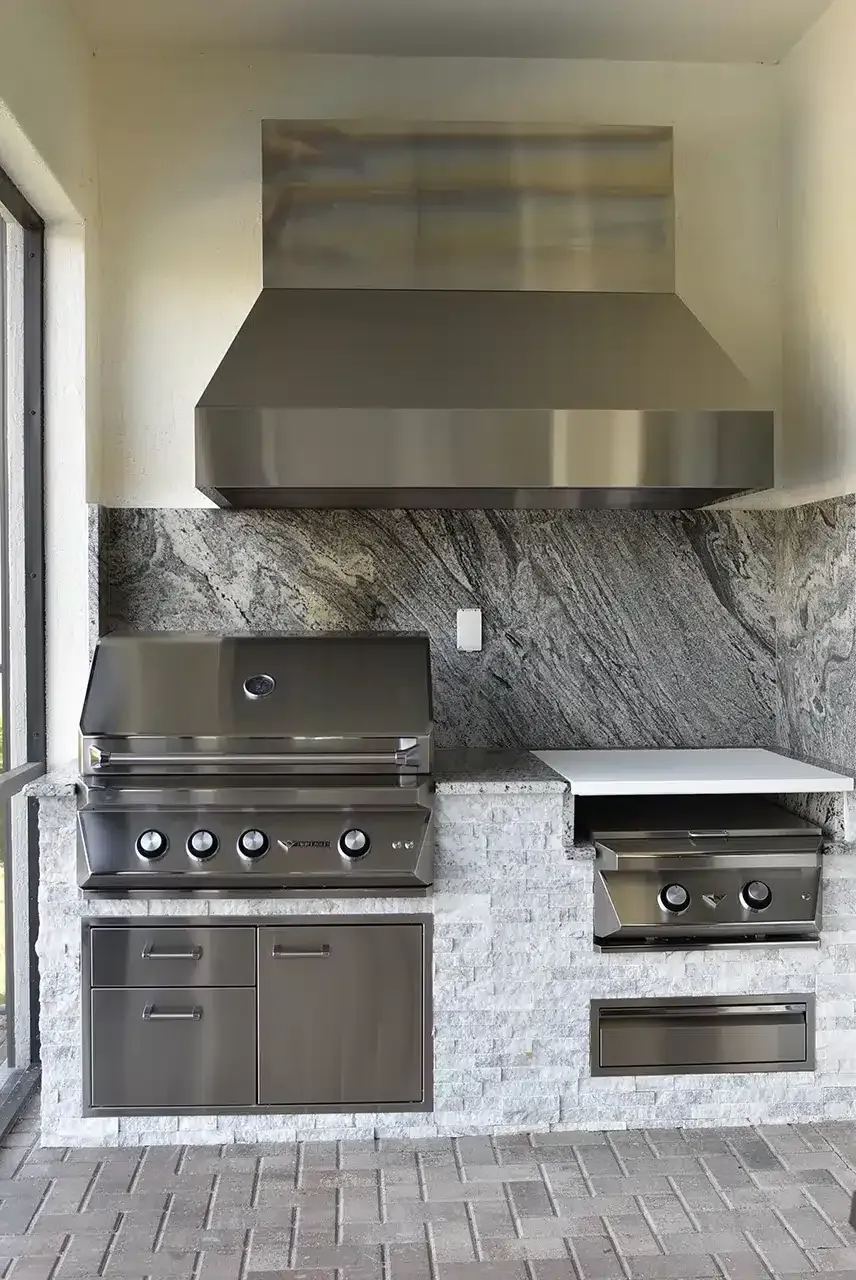 Outdoor stainless steel kitchen with built-in grill, side burner, and hood against a stone backsplash