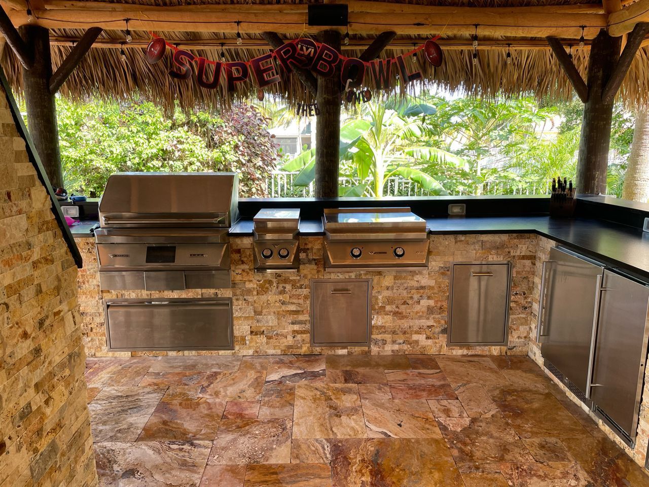 Covered outdoor kitchen with stainless steel grill, sink, and cabinets under a thatched roof