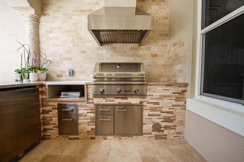 Modern outdoor kitchen with stainless steel grill, sink, cabinets, and beige tile backsplash