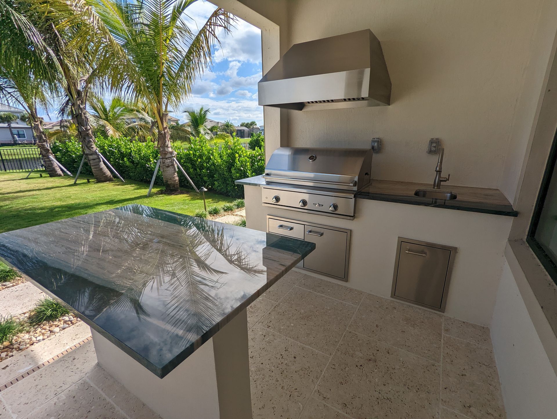 Outdoor patio kitchen with grill, sink, and stone countertop beside a tropical garden