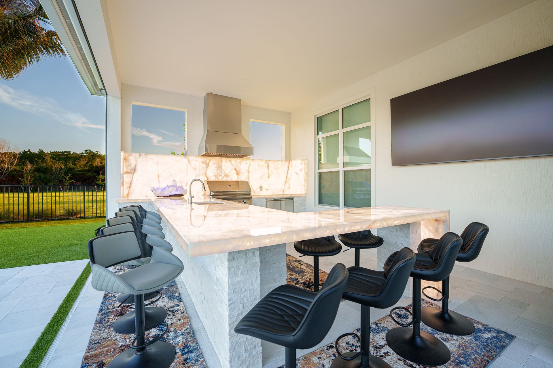 Modern outdoor kitchen with bar seating, marble island, grill, and wall-mounted TV under a covered patio