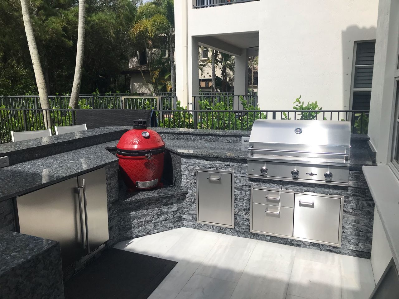 Outdoor patio with stainless steel grill, red smoker, and dark granite countertops beside a white house.
