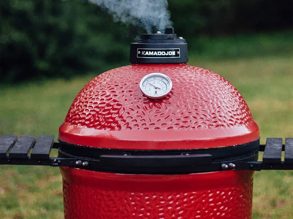Red ceramic smoker grill with thermometer and smoke rising from the top vent.