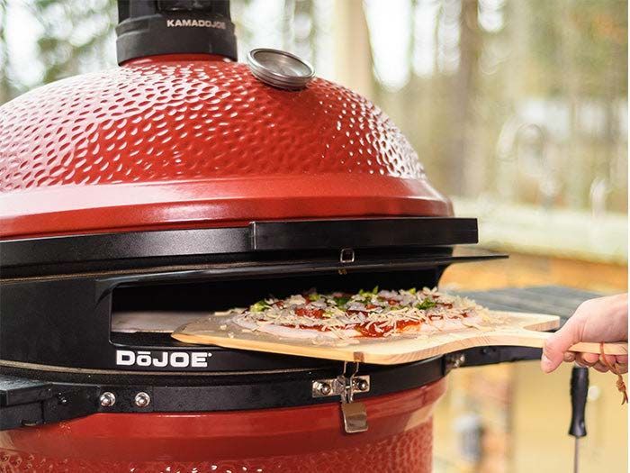 Pizza being pulled from a red domed outdoor grill with a wooden peel
