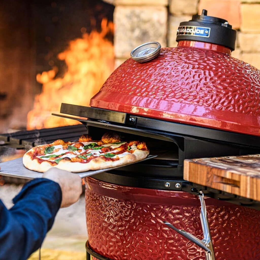 Person sliding a pizza into a red ceramic outdoor pizza oven with a fire burning inside