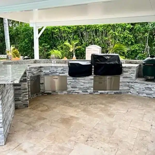 Covered outdoor patio with stone kitchen counters, two grills, and a tiled floor surrounded by greenery