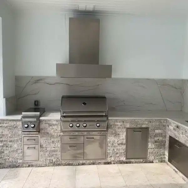 Outdoor kitchen with built-in stainless steel grill, hood, counter, and storage cabinets against a stone wall.