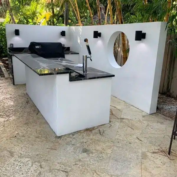 Outdoor white kitchen island with black countertop, sink, grill, and tall white privacy wall.