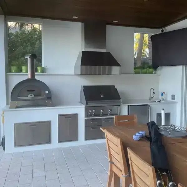 Outdoor kitchen with pizza oven, grill, sink, and wood dining table under a covered patio