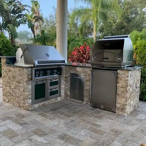 Outdoor stone barbecue and grill station with two stainless steel grills on a patio, surrounded by palm trees.