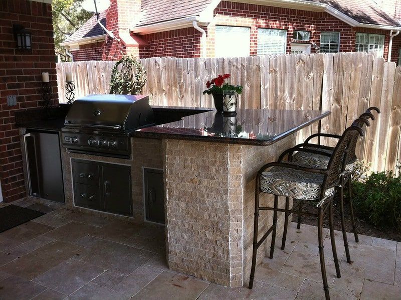 Covered patio with built-in grill, granite counter, and two bar stools beside a wooden fence.