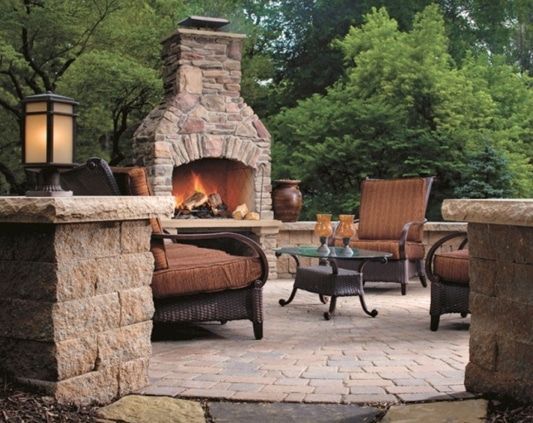 Stone patio with outdoor fireplace, wicker chairs, and lanterns surrounded by lush greenery.