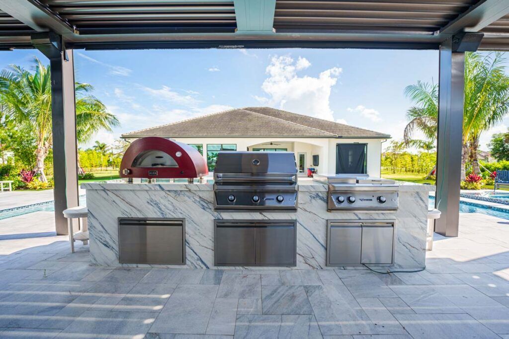 Outdoor kitchen with grill, pizza oven, and sink under a covered patio by a pool.