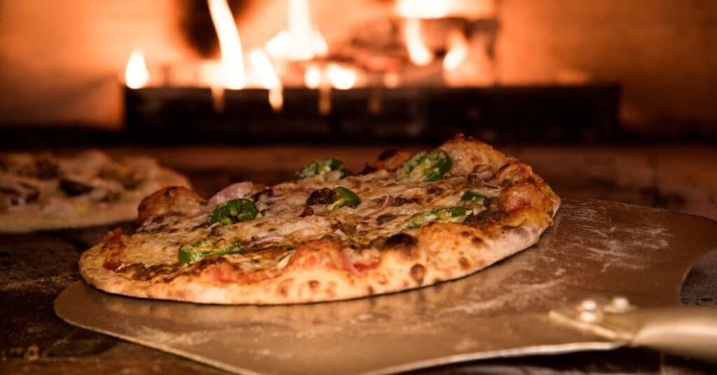 Pizza baking in a wood-fired oven, with a pizza peel in the foreground and warm flames behind.