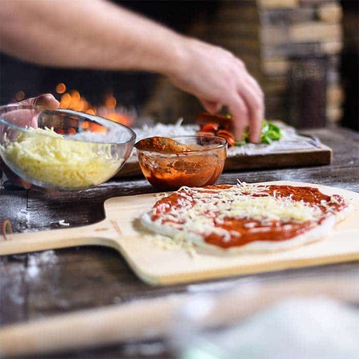 Pizza being assembled on a wooden board, with bowls of cheese, sauce, and toppings on a kitchen counter