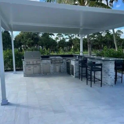 Outdoor patio with a covered stone bar, grill, and black stools on a tiled terrace surrounded by greenery