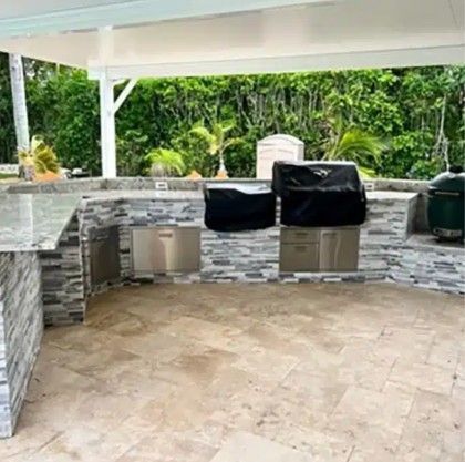Covered outdoor stone kitchen with grill, sink, and lush green garden view