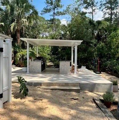 Outdoor patio with a white pergola, concrete seating, and surrounding tropical trees and plants