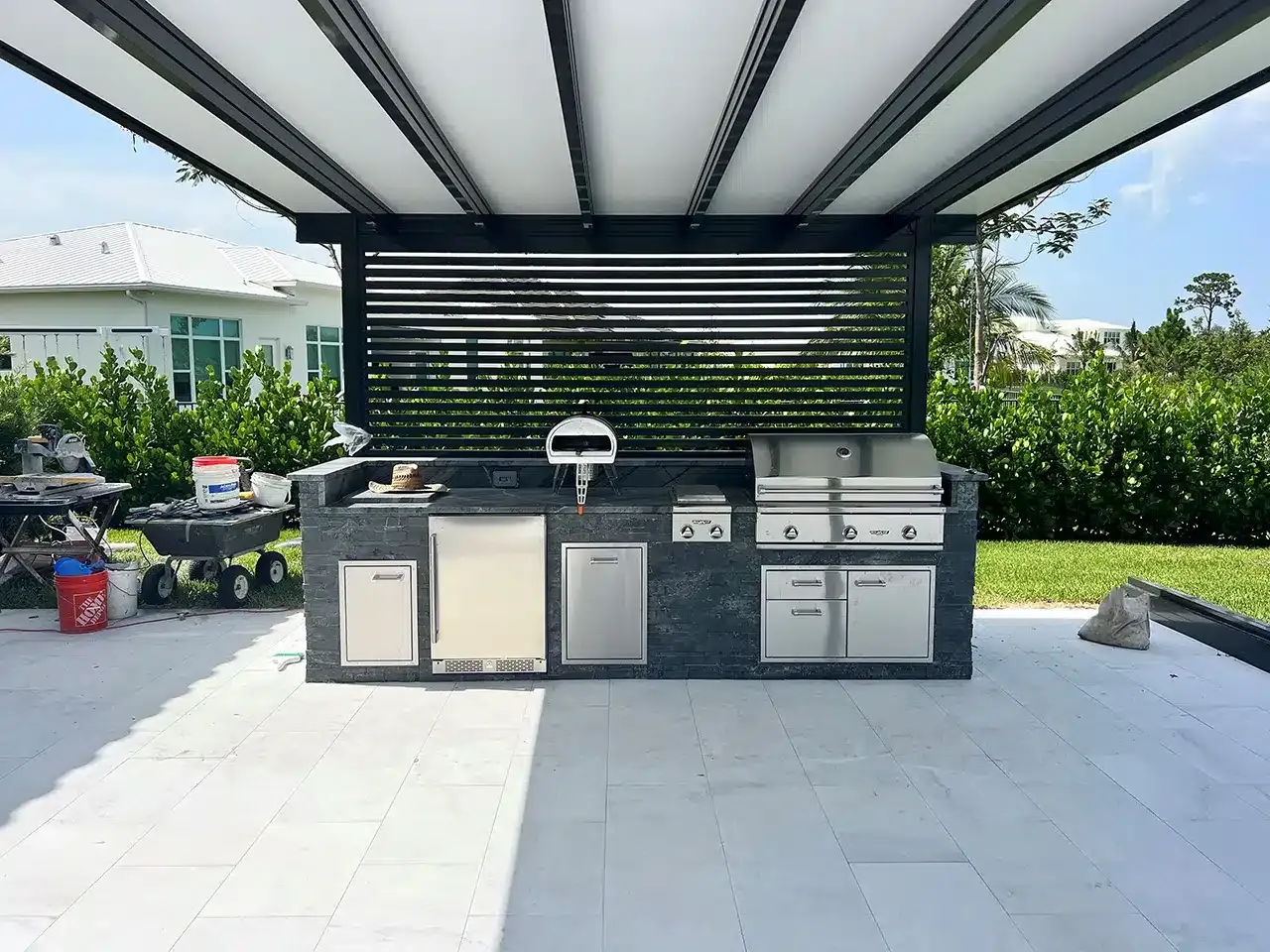 Outdoor patio kitchen with grill, sink, cabinets, and black slatted backsplash under a covered pergola