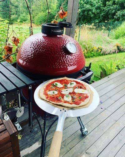 Fresh pizza on a peel beside a red dome grill on a wooden deck overlooking a green garden
