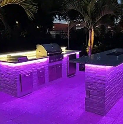 Purple-lit outdoor kitchen with a built-in grill and stone counters under palm trees at night