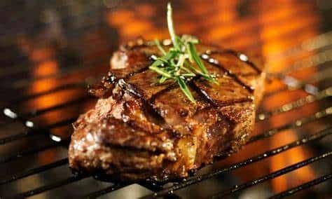 Grilled steak on a barbecue grate, topped with fresh herbs and glowing with heat from the flames behind it