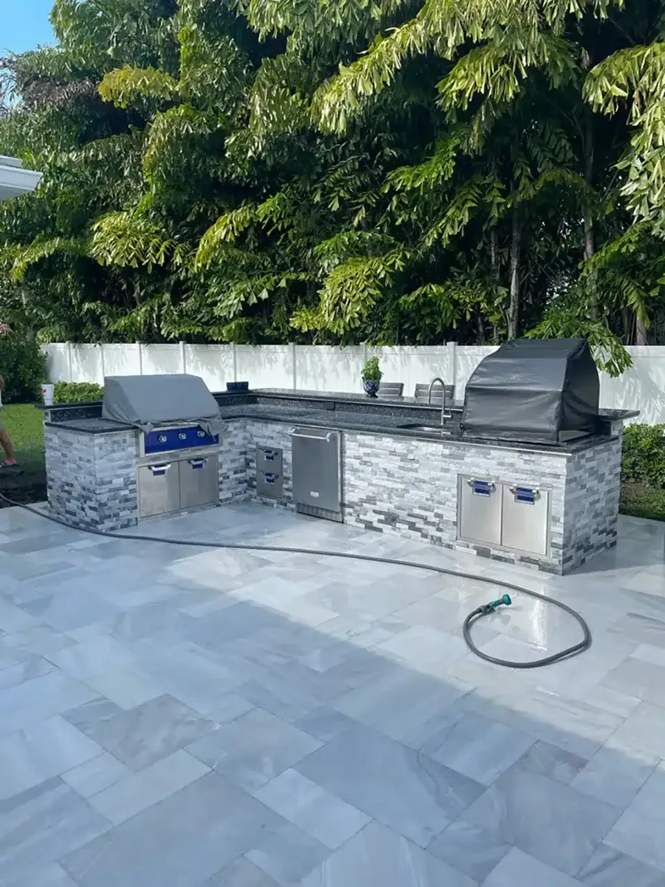 Outdoor stone patio with built-in grill, sink, and black grill cover under a large tree
