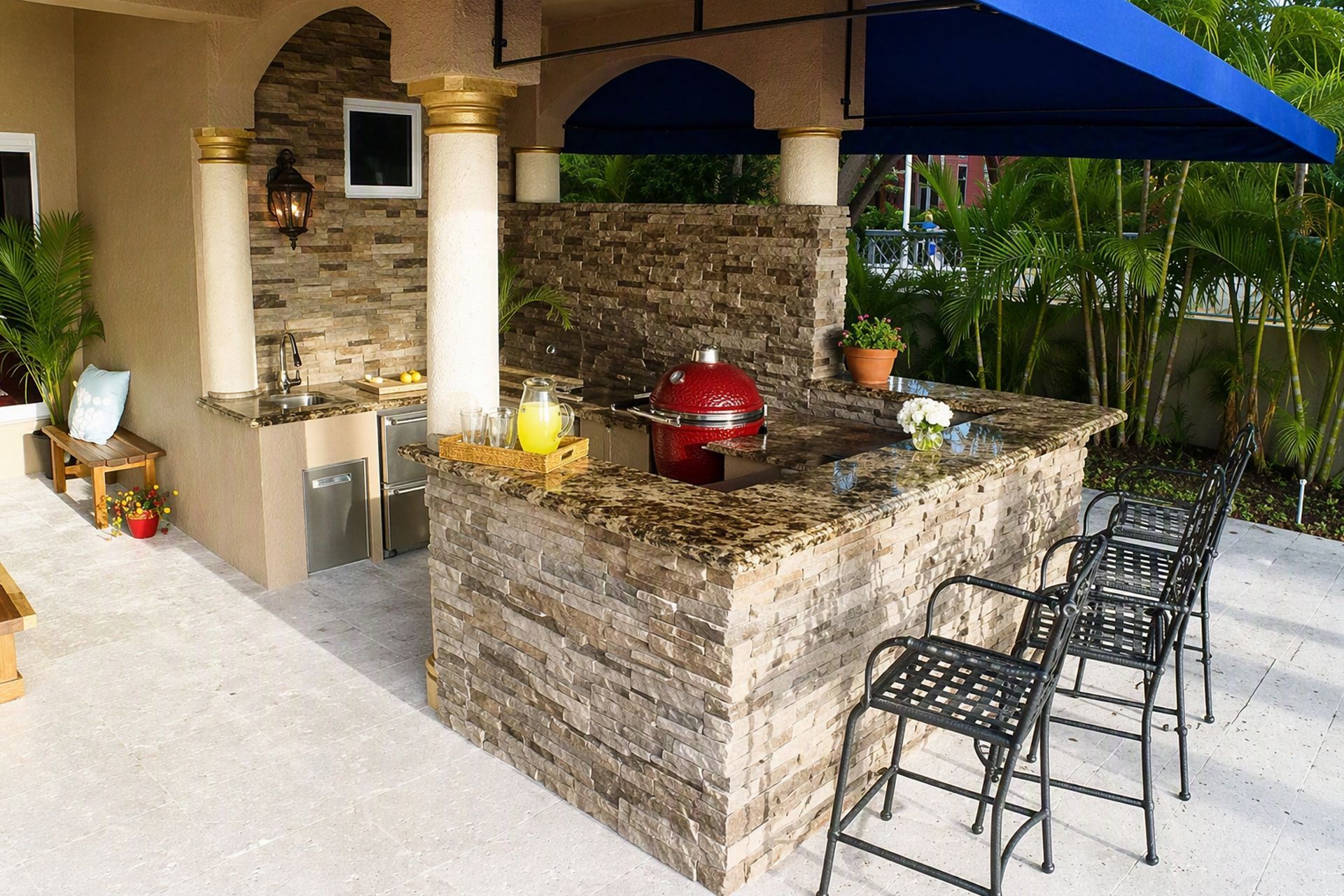 Outdoor stone patio with built-in grill, bar counter, and black bar stools under a blue awning