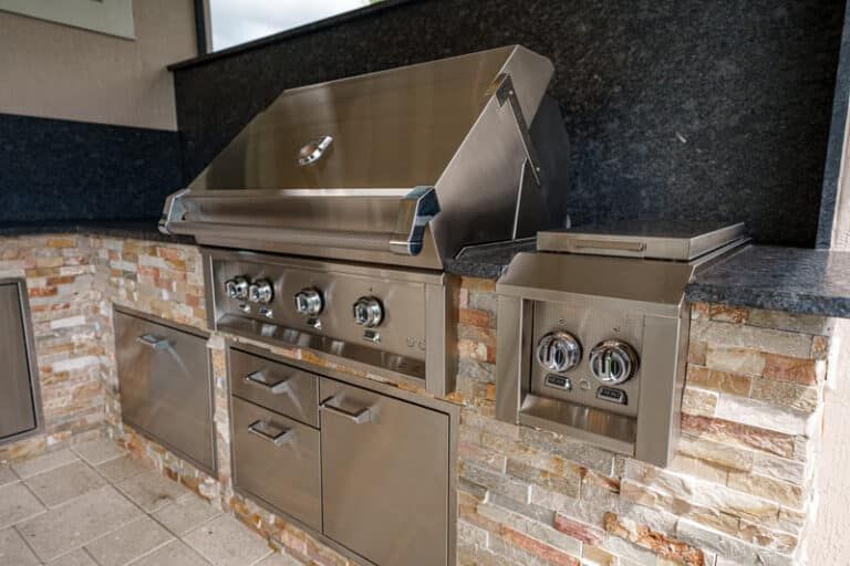 Outdoor built-in grill station with red pizza oven, stainless barbecue, and sink under a shaded patio.