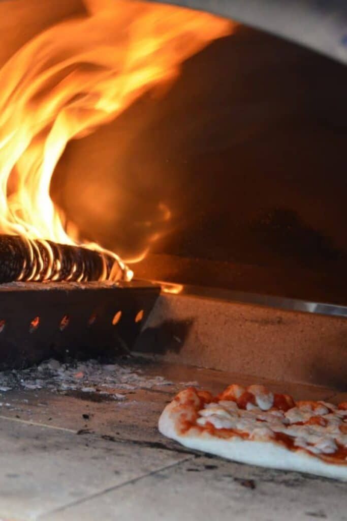 Pizza baking in a wood-fired oven, with bright flames and a partially cooked pie in the foreground