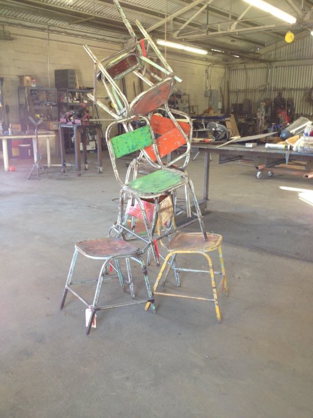 Chairs stacked precariously in a workshop. Several stools at the base. Colors include red, green, and yellow.