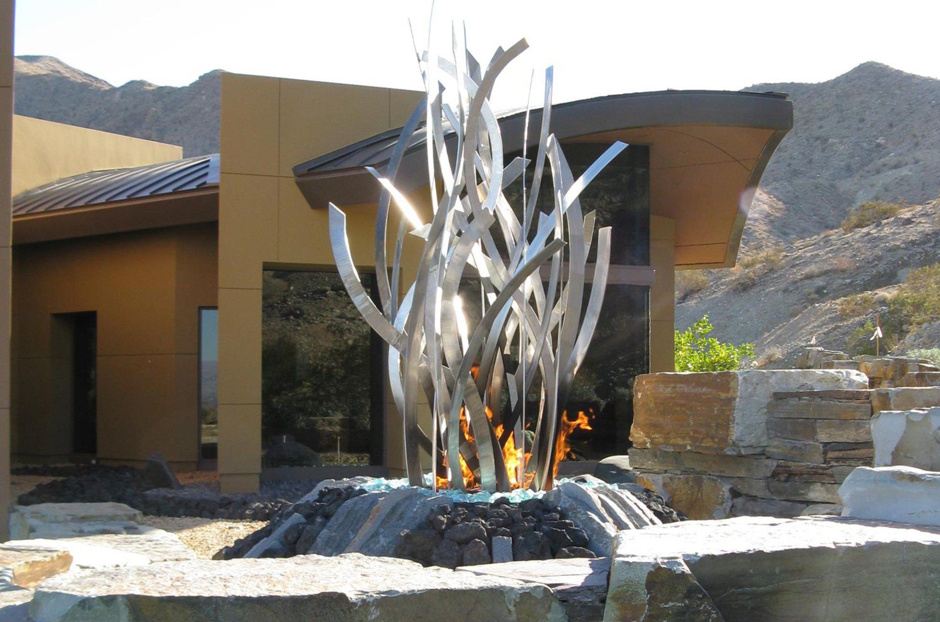 Outdoor fire feature with metal sculpture, flames, and modern building.