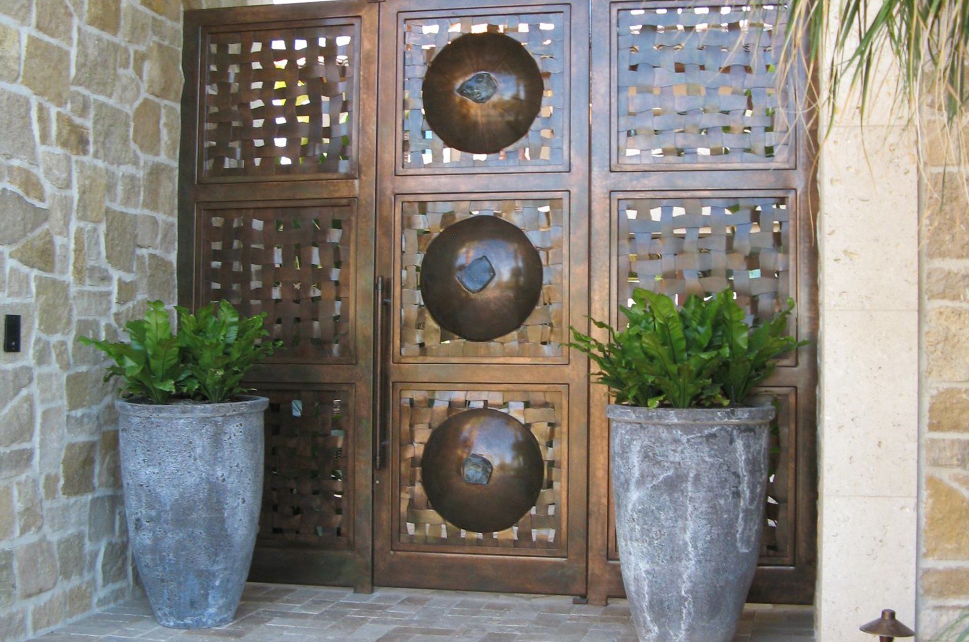 Stone-walled entrance with bronze metal doors, large pots, and green plants.