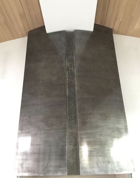 Metal range hood with a decorative center strip, against a white wall and wood ceiling.