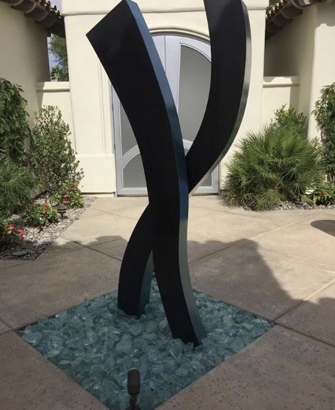 Black abstract sculpture on a bed of green glass rocks, in front of a white building with arched doors.