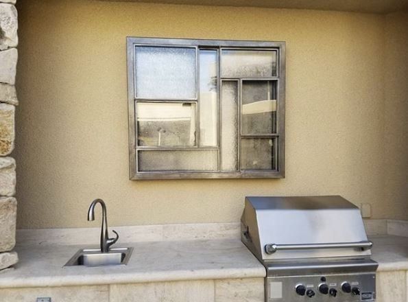 Outdoor kitchen with a stainless steel grill, sink, and window against a tan wall.