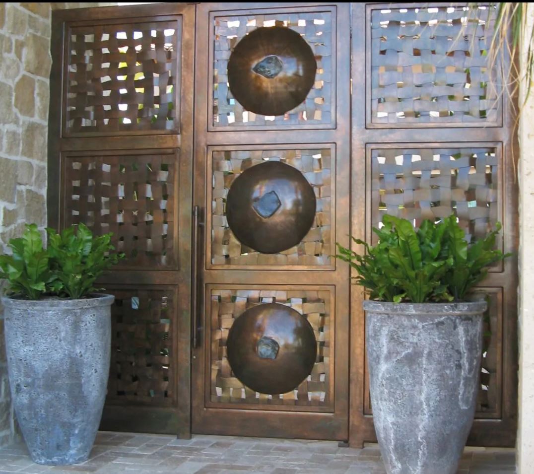 Bronze gate with woven design and three large circular accents, flanked by potted plants.