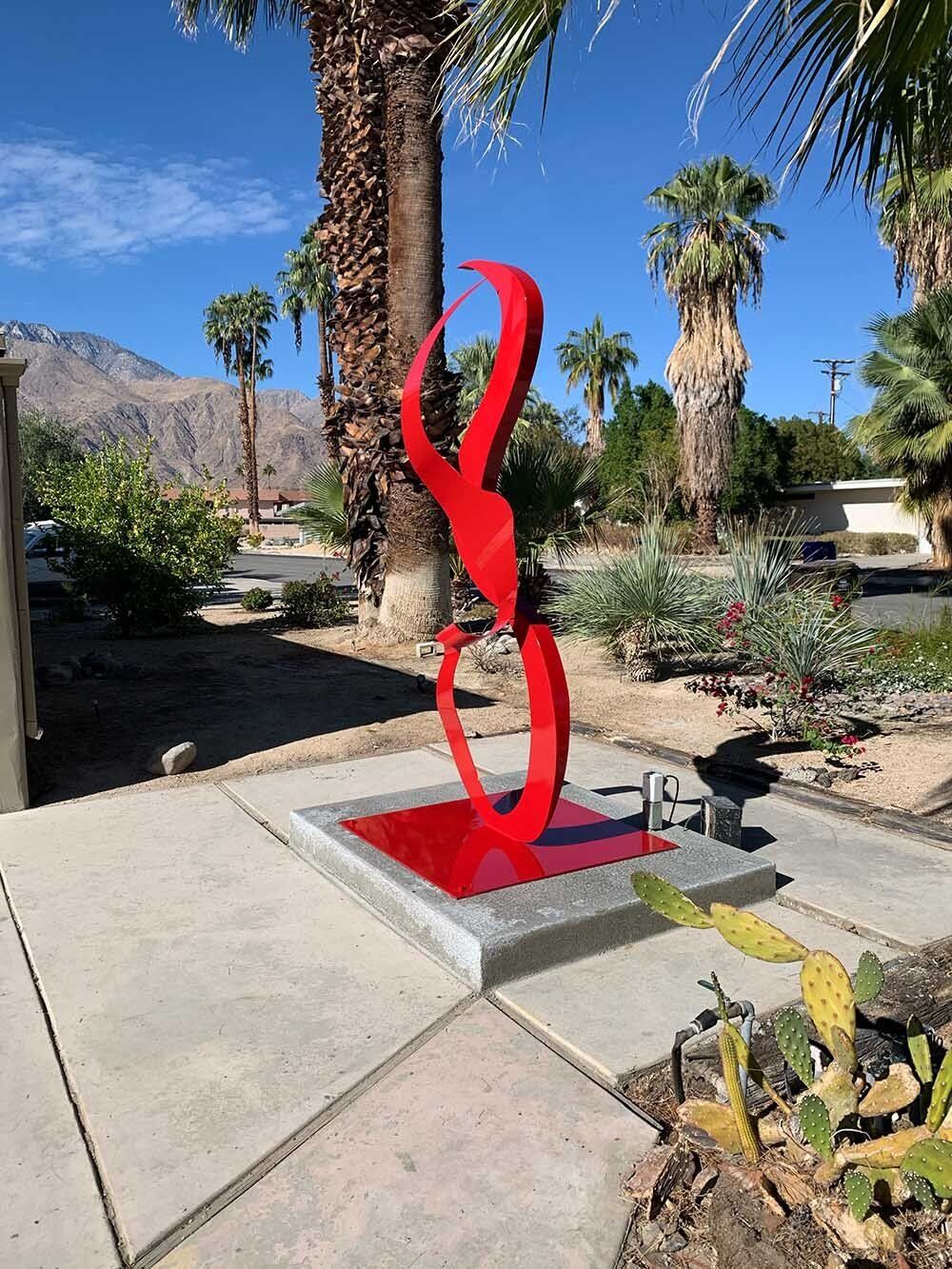 Red abstract sculpture on a concrete base in a sunny outdoor setting with palm trees and mountains.
