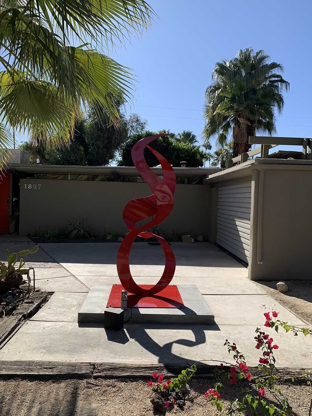 Red metal sculpture in garden, with palm trees and stucco wall in background.