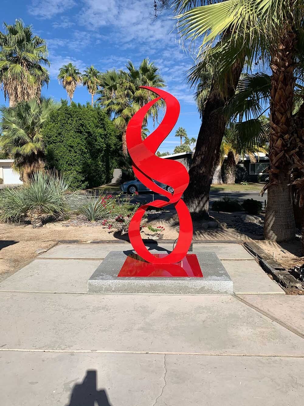 Red abstract sculpture on a concrete base, framed by palms, in an outdoor setting.
