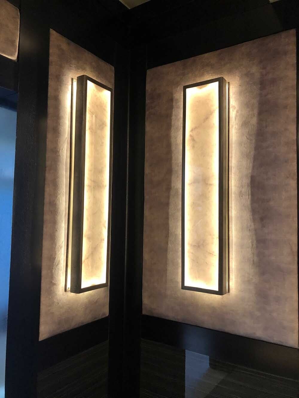 Two rectangular, backlit wall sconces in a dark corner, mounted on textured, pale pink panels.