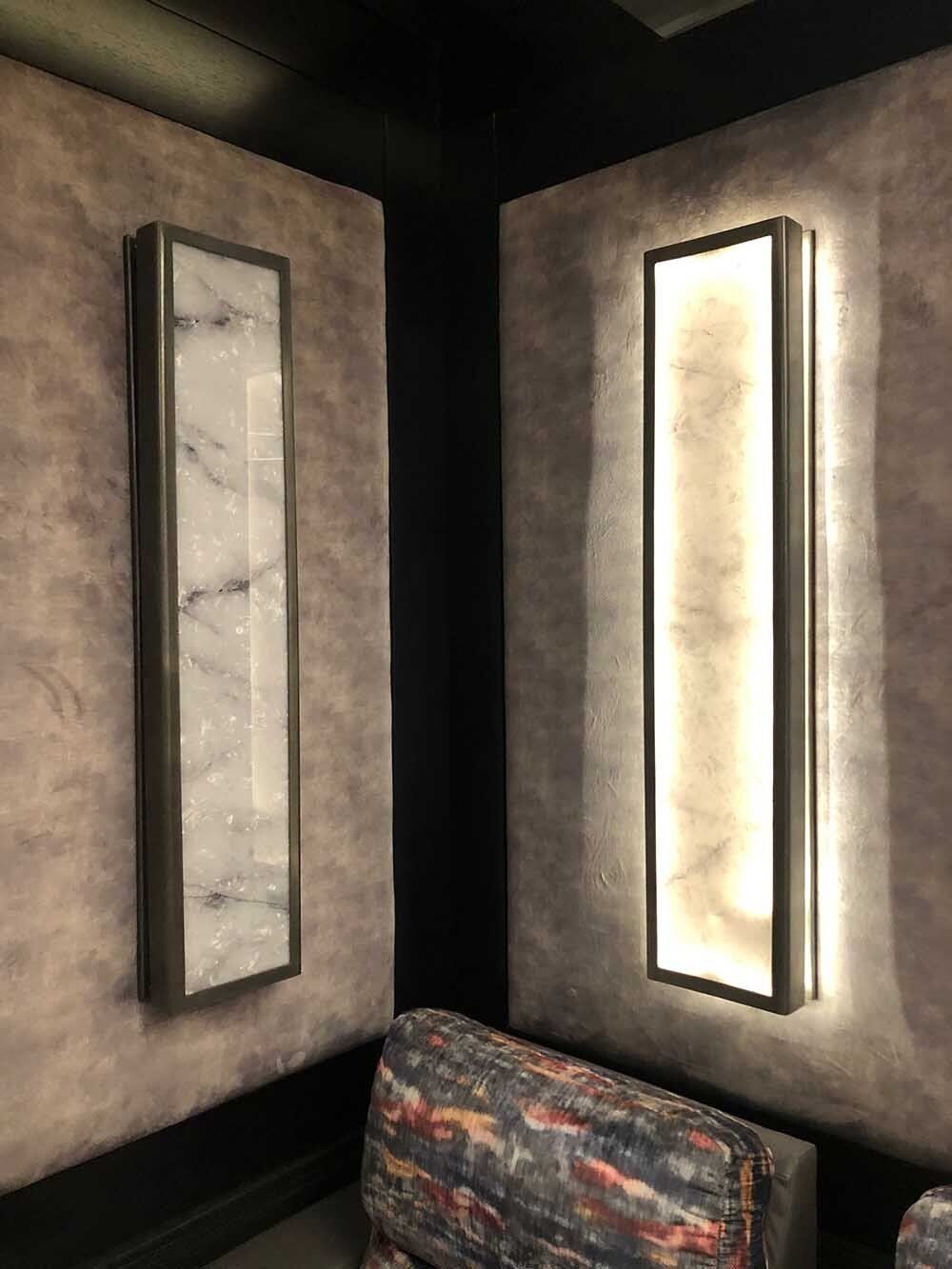 Two rectangular wall sconces, one marble, one illuminated, in a corner with upholstered walls.
