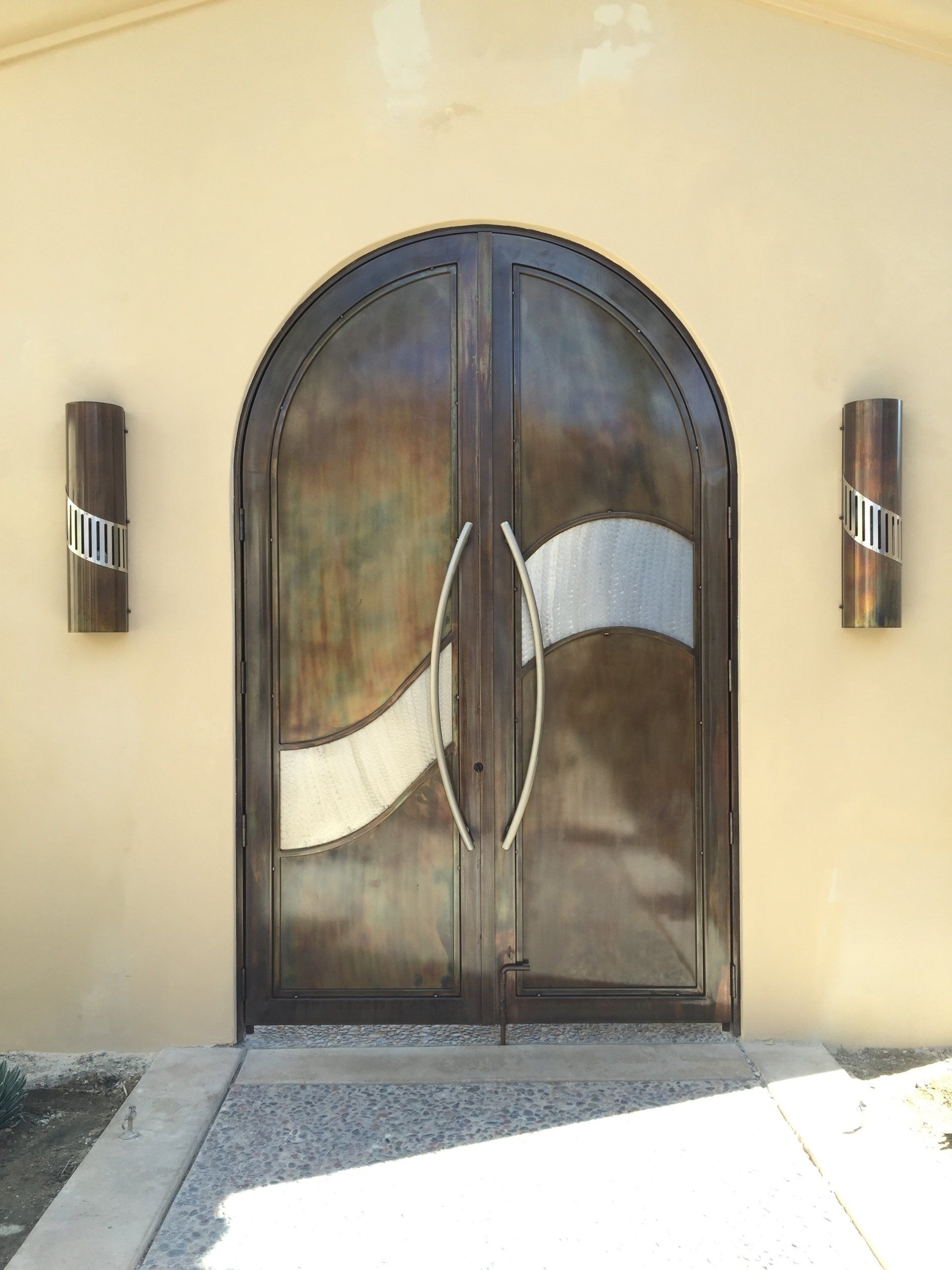 Arched bronze double doors with a silver accent, flanked by cylindrical sconces, set in a beige wall.